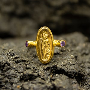 Könnte beinhalten: Ein goldener Ring mit einem erhabenen ovalen Design, das eine Frau in einem fließenden Gewand zeigt. Der Ring hat zwei kleine violette Edelsteine ​​auf beiden Seiten des Ovals.