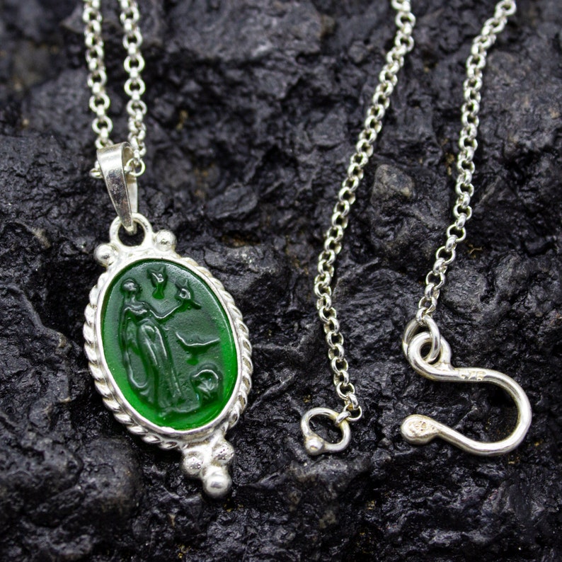 Puede incluir: Un collar de cadena de plata con un colgante de vidrio verde que presenta una imagen tallada de una mujer sosteniendo un p&aacute;jaro. El colgante tiene forma ovalada y tiene un borde de cuerda.