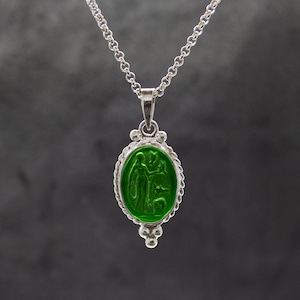 Puede incluir: Un collar de cadena de plata con un colgante ovalado verde que presenta un dise&ntilde;o en relieve de dos figuras. El colgante est&aacute; rodeado por un borde plateado con un dise&ntilde;o de cuerda.