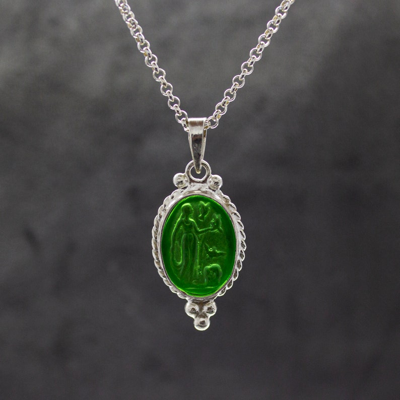 Puede incluir: Un collar de cadena de plata con un colgante ovalado verde que presenta un dise&ntilde;o en relieve de dos figuras. El colgante est&aacute; rodeado por un borde plateado con un dise&ntilde;o de cuerda.