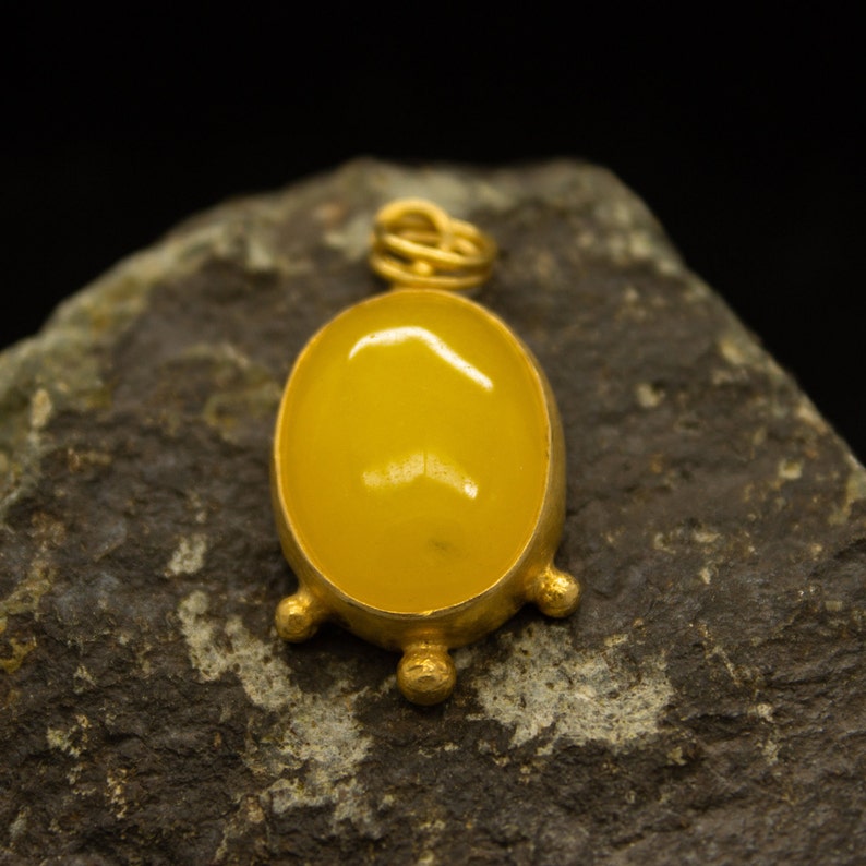 May include: A gold pendant with a yellow oval gemstone set in a bezel setting. The pendant has a decorative bail and four small gold balls on the bottom.