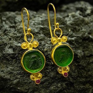 May include: Gold dangle earrings with green glass insets featuring a carved design. The earrings have a red gemstone accent.