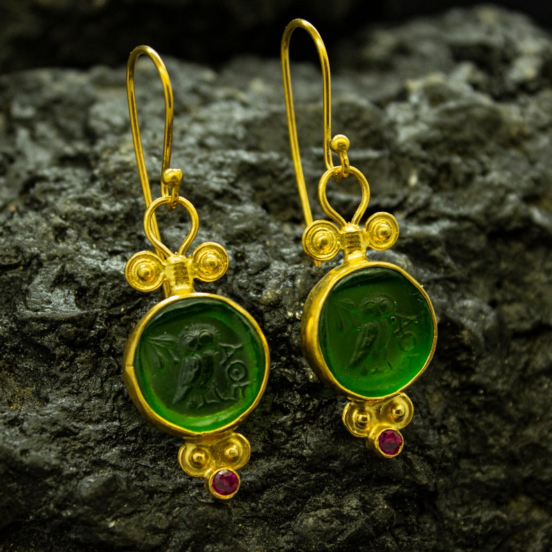 May include: Gold dangle earrings with green glass insets featuring a carved design. The earrings have a red gemstone accent.