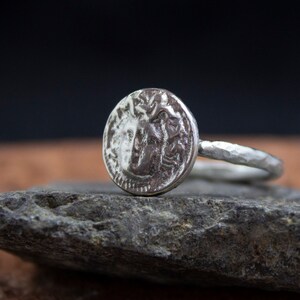 Op de afbeelding: Een zilveren ring met een getextureerde band en een ronde, muntvormige bovenkant met een gedetailleerd ontwerp.
