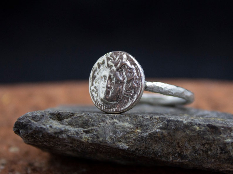 Op de afbeelding: Een zilveren ring met een getextureerde band en een ronde, muntvormige bovenkant met een gedetailleerd ontwerp.