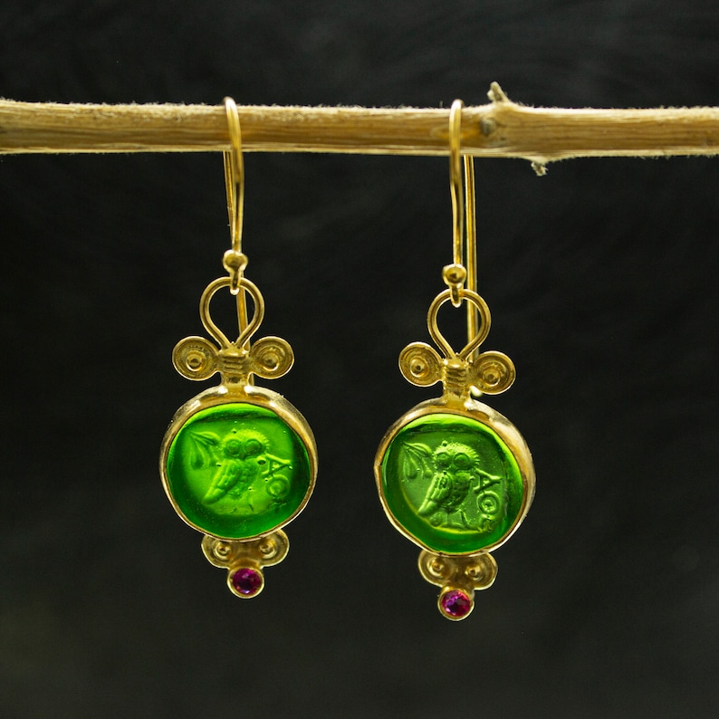 May include: Gold earrings with green glass owl charms and pink gemstones. The owls are in a cameo-like design.