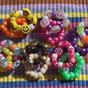May include: A collection of colorful beaded bracelets with smiley face and flower charms. The bracelets are made of plastic beads and are stacked on a striped fabric background.