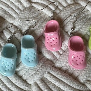 May include: Six pairs of colorful dangle earrings shaped like clogs. The earrings are made of ceramic and have a glossy finish. The colors include light purple, light blue, pink, and green.