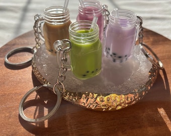 May include: Four miniature glass jars with clear lids and straws, filled with different colored liquids, resembling bubble tea. The jars are attached to silver keychains and are displayed on a gold and white decorative surface.
