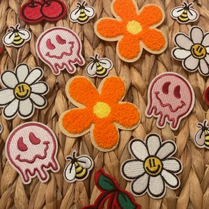 May include: A collection of colorful iron-on patches featuring various designs, including smiley faces, flowers, cherries, and bees. The patches are arranged on a woven wicker surface.