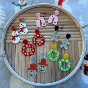 May include: A collection of Christmas-themed earrings featuring a snowman, wreath, gingerbread man, and candy cane designs. The earrings are made of polymer clay and are painted in various colors, including red, green, white, and pink.