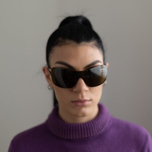 May include: A woman with dark hair pulled back in a ponytail is wearing a purple turtleneck sweater and dark sunglasses.