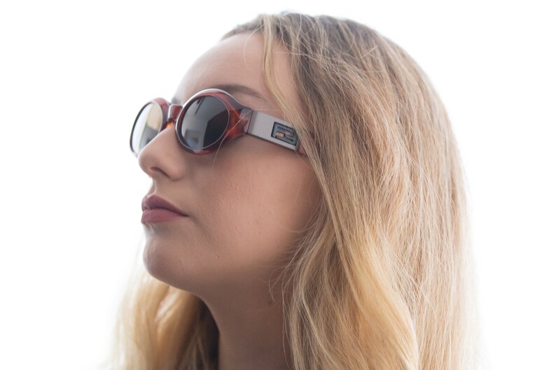 May include: A woman wearing brown and silver oval sunglasses with a reflective lens. The sunglasses have a silver frame and a brown tortoise shell pattern.