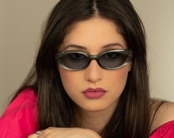 May include: A woman with long dark hair wearing oval-shaped sunglasses with a dark gray frame and lenses. She is wearing a bright pink off-the-shoulder top.
