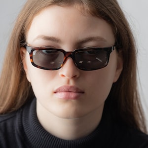 Könnte beinhalten: Eine junge Person mit langen braunen Haaren, die einen schwarzen Rollkragenpullover und eine Schildpatt-Sonnenbrille trägt.