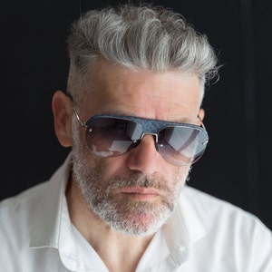 Op de afbeelding: Een man met grijs haar en een baard die een wit overhemd en een blauwe zonnebril draagt.