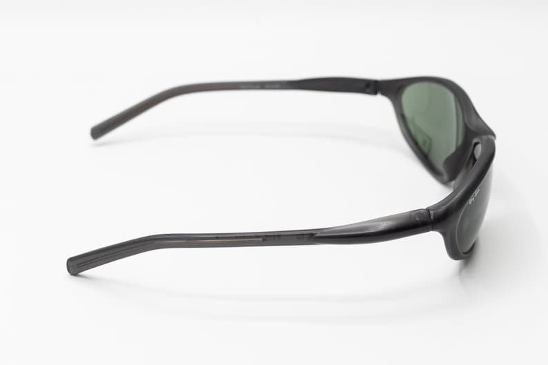 May include: Black sunglasses with green lenses. The sunglasses have a curved frame and a thin, flexible arm.