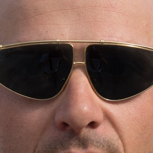 Könnte beinhalten: Nahaufnahme einer Person, die eine goldene Sonnenbrille mit schwarzen Gläsern trägt. Die Sonnenbrille hat eine einzigartige, eckige Form.