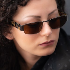 May include: Rectangular sunglasses with a dark metal frame and brown lenses. The temples feature a tortoiseshell pattern. The image is a close-up, focusing on the sunglasses and the person wearing them.