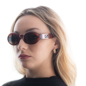 May include: A woman wearing brown oval sunglasses with a silver Fendi logo on the side. The sunglasses have dark lenses and a tortoise shell frame.