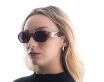 May include: A woman wearing brown oval sunglasses with a silver Fendi logo on the side. The sunglasses have dark lenses and a tortoise shell frame.
