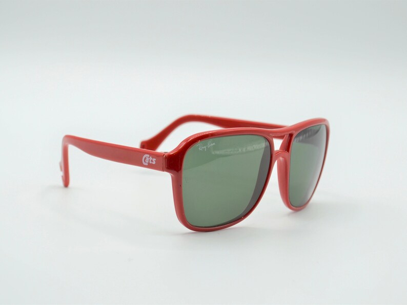 May include: Red Ray-Ban sunglasses with green lenses. The sunglasses have a white "Cats" logo on the side.