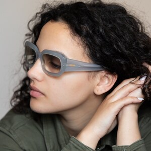 May include: A woman with curly dark hair wearing gray square-shaped sunglasses with a gold accent.