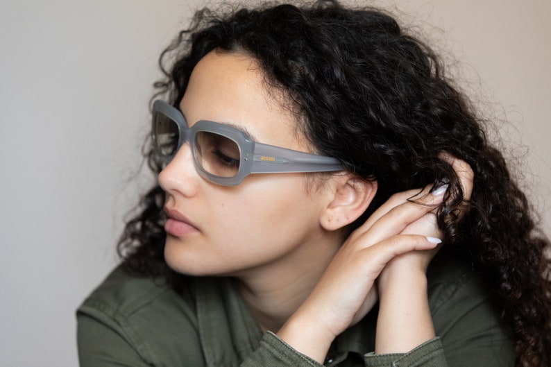 May include: A woman with curly dark hair wearing gray square-shaped sunglasses with a gold accent.