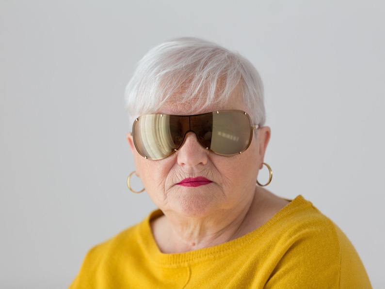 May include: A woman with short, white hair wearing large, gold-rimmed sunglasses and gold hoop earrings. She is wearing a yellow sweater.