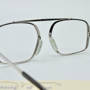 Puede incluir: Gafas de metal plateadas con montura rectangular y lentes transparentes. Las gafas tienen una bisagra de resorte y almohadillas nasales transparentes.