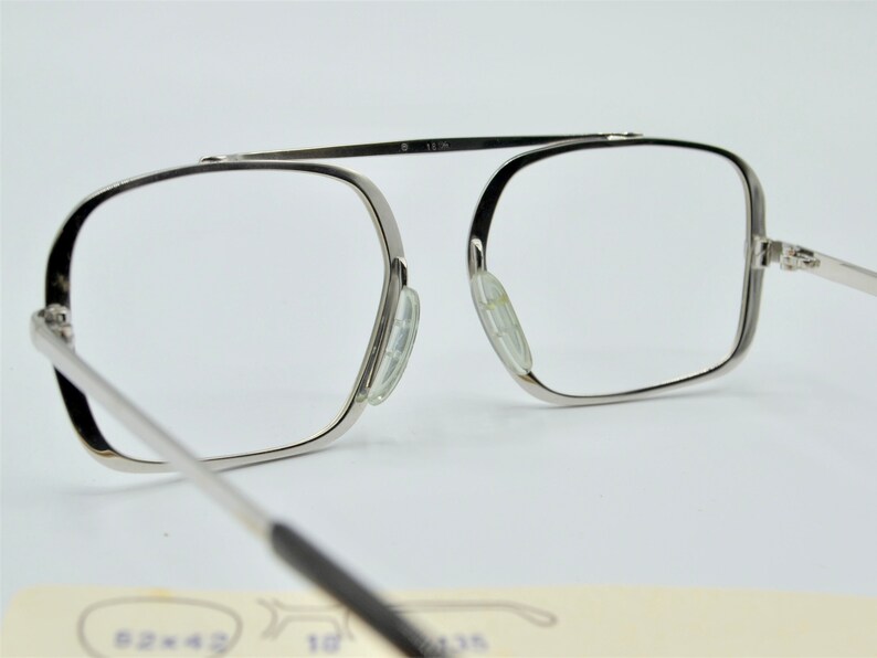 Puede incluir: Gafas de metal plateadas con montura rectangular y lentes transparentes. Las gafas tienen una bisagra de resorte y almohadillas nasales transparentes.