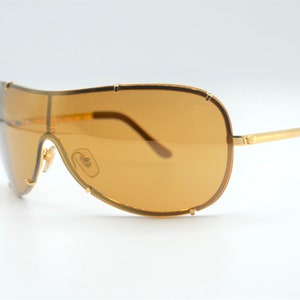 May include: Gold-tone aviator sunglasses with brown lenses. The sunglasses have a unique, curved frame design.