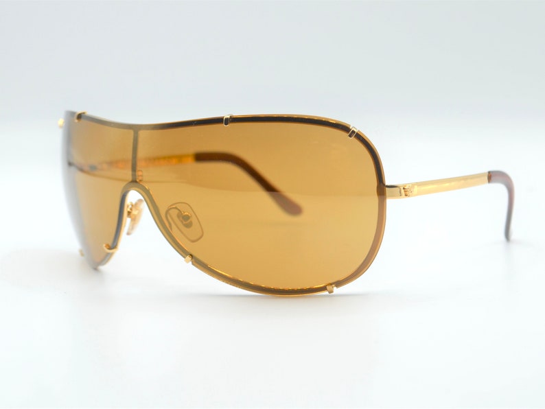 May include: Gold-tone aviator sunglasses with brown lenses. The sunglasses have a unique, curved frame design.