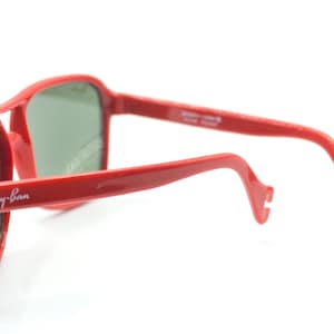 May include: Red Ray-Ban sunglasses with green lenses. The sunglasses have a classic aviator style with a thin metal frame and a curved bridge.