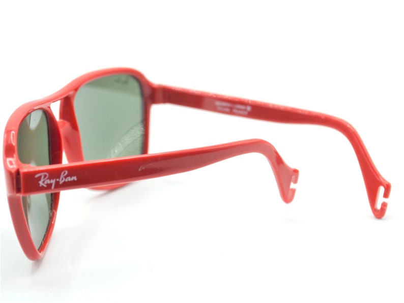 May include: Red Ray-Ban sunglasses with green lenses. The sunglasses have a classic aviator style with a thin metal frame and a curved bridge.