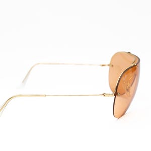 K&ouml;nnte beinhalten: Sonnenbrille mit goldfarbenem Rahmen und hellbraunen Gl&auml;sern. Die Brille im Aviator-Stil hat einen d&uuml;nnen Goldrahmen und ein geschwungenes Linsendesign. Die B&uuml;gel sind ebenfalls goldfarben, mit transparenten Kunststoff-Ohrb&uuml;geln.