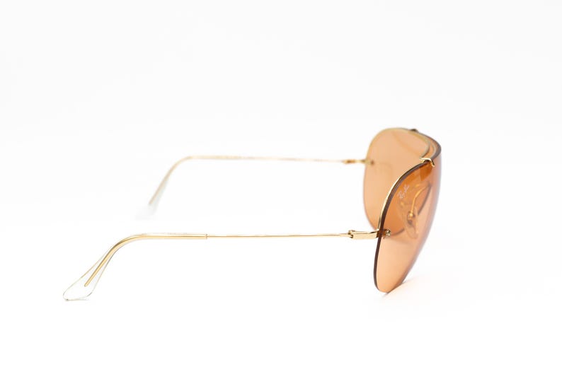 K&ouml;nnte beinhalten: Sonnenbrille mit goldfarbenem Rahmen und hellbraunen Gl&auml;sern. Die Brille im Aviator-Stil hat einen d&uuml;nnen Goldrahmen und ein geschwungenes Linsendesign. Die B&uuml;gel sind ebenfalls goldfarben, mit transparenten Kunststoff-Ohrb&uuml;geln.
