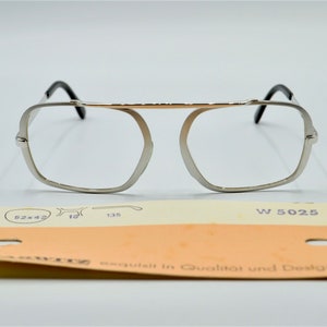 Puede incluir: Gafas de metal plateadas con puente dorado y patillas negras. Las gafas tienen una forma rectangular con esquinas redondeadas. La etiqueta en la parte inferior dice "W 5025" y "exquisit in Qualitat und Design".