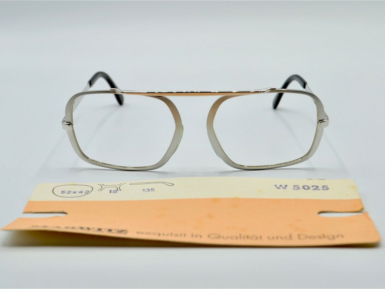 Puede incluir: Gafas de metal plateadas con puente dorado y patillas negras. Las gafas tienen una forma rectangular con esquinas redondeadas. La etiqueta en la parte inferior dice "W 5025" y "exquisit in Qualitat und Design".