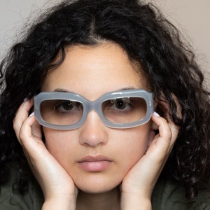 May include: A person with curly dark hair wearing gray tinted sunglasses. The person is looking down with their hands resting on their cheeks.