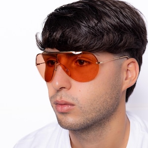K&ouml;nnte beinhalten: Orange get&ouml;nte Pilotenbrille mit goldfarbenem Metallrahmen. Die Sonnenbrille hat einen Doppelsteg und einen gebogenen oberen B&uuml;gel. Die Gl&auml;ser sind einfarbig orange und die B&uuml;gel sind d&uuml;nn und goldfarben. Getragen von einer Person.
