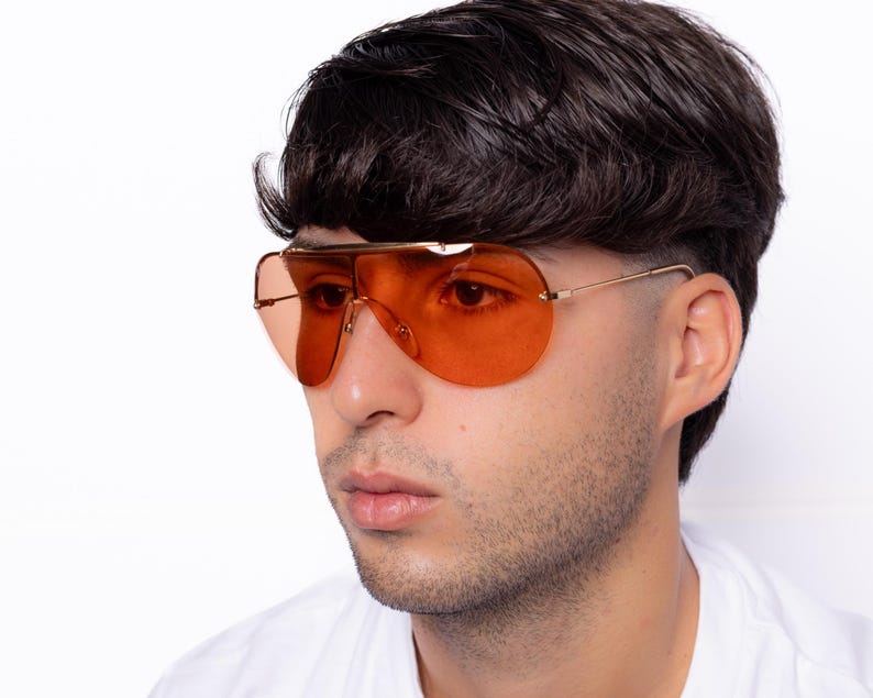 K&ouml;nnte beinhalten: Orange get&ouml;nte Pilotenbrille mit goldfarbenem Metallrahmen. Die Sonnenbrille hat einen Doppelsteg und einen gebogenen oberen B&uuml;gel. Die Gl&auml;ser sind einfarbig orange und die B&uuml;gel sind d&uuml;nn und goldfarben. Getragen von einer Person.