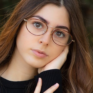 Peut inclure: Une jeune personne aux longs cheveux bruns portant des lunettes rondes à monture brune avec le mot "VIENNA" sur la lentille droite. La personne porte un pull noir et a la main sur le menton.