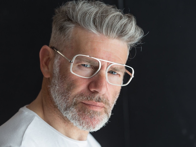 Puede incluir: Un hombre con cabello gris y barba lleva gafas con montura blanca y puente de metal plateado. Lleva una camiseta blanca.