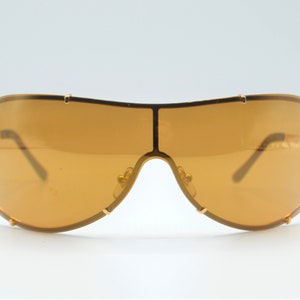 May include: Gold-tone aviator sunglasses with brown lenses. The sunglasses have a wraparound design and a thick frame.