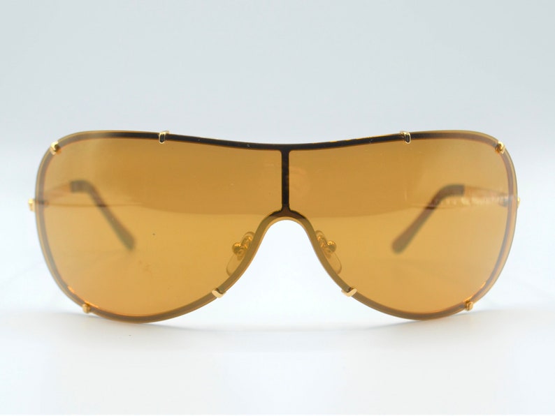 May include: Gold-tone aviator sunglasses with brown lenses. The sunglasses have a wraparound design and a thick frame.