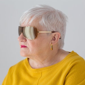 May include: A woman with short, white hair wearing large, brown, mirrored sunglasses and a yellow sweater.
