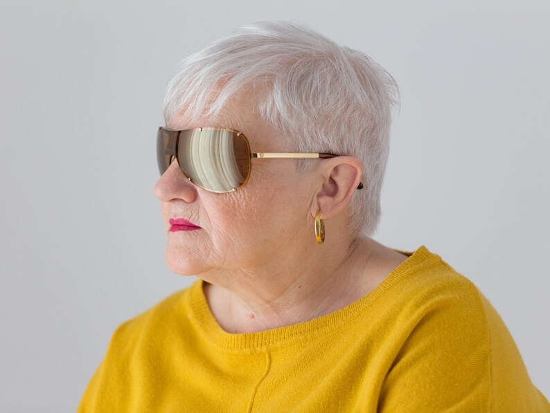 May include: A woman with short, white hair wearing large, brown, mirrored sunglasses and a yellow sweater.