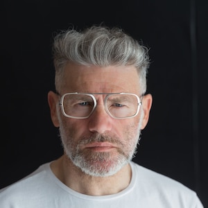 Puede incluir: Un hombre con cabello gris y barba lleva gafas con montura plateada. Est&aacute; mirando directamente a la c&aacute;mara con una expresi&oacute;n seria.