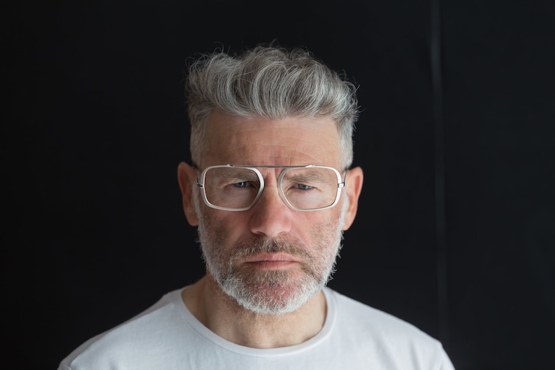 Puede incluir: Un hombre con cabello gris y barba lleva gafas con montura plateada. Est&aacute; mirando directamente a la c&aacute;mara con una expresi&oacute;n seria.